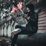 A person wearing a mask reads a flaming newspaper on an urban street, creating a surreal and mysterious scene.